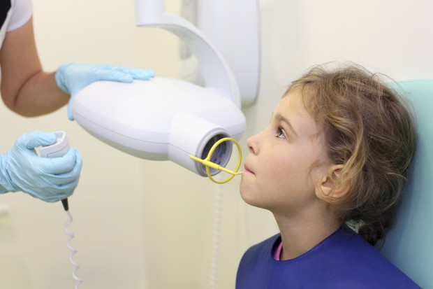 A little girl sitting in a chair, wearing a lead apron as the dentist performs an intraoral X-ray. An extraoral X-ray is another way of looking at your teeth.